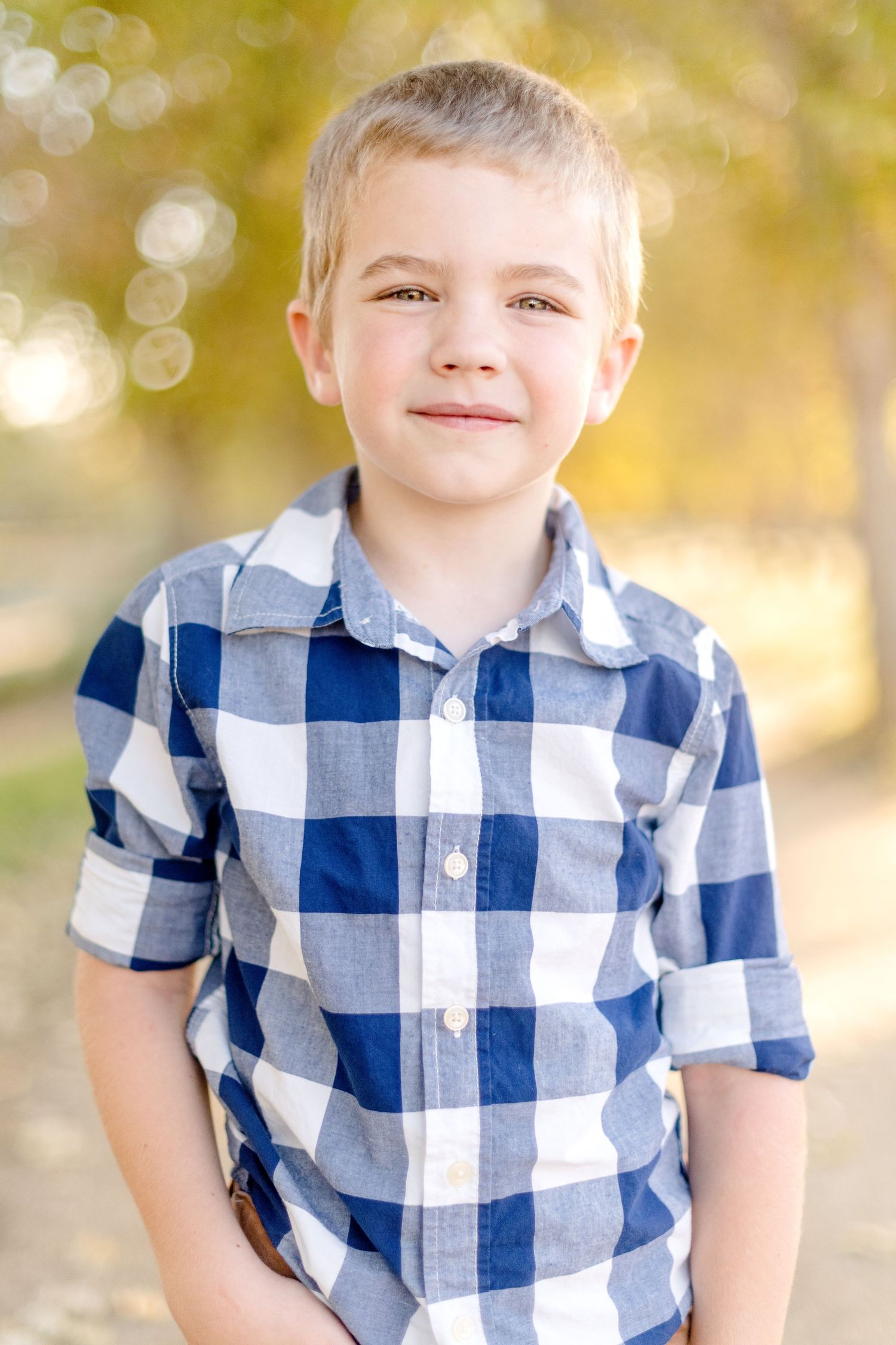 Young child wearing a blue and white checkered button-down shirt smiles outdoors in autumn lighting.