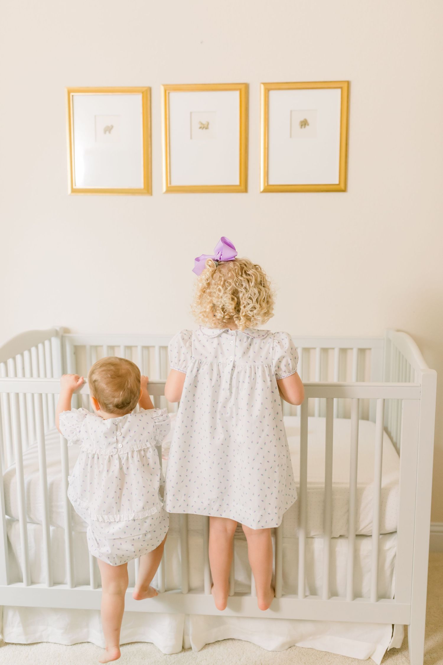 Two small children in light dresses peer into a white crib beneath golden frames on the wall.