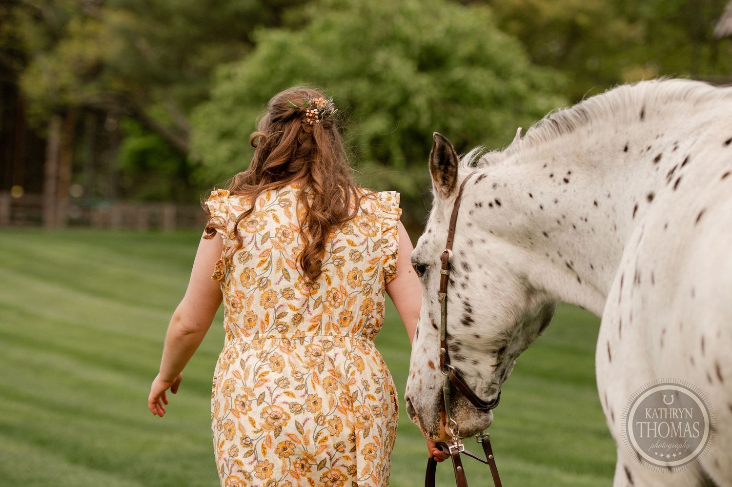 A spotted white horse follows behind someone wearing a yellow floral dress.