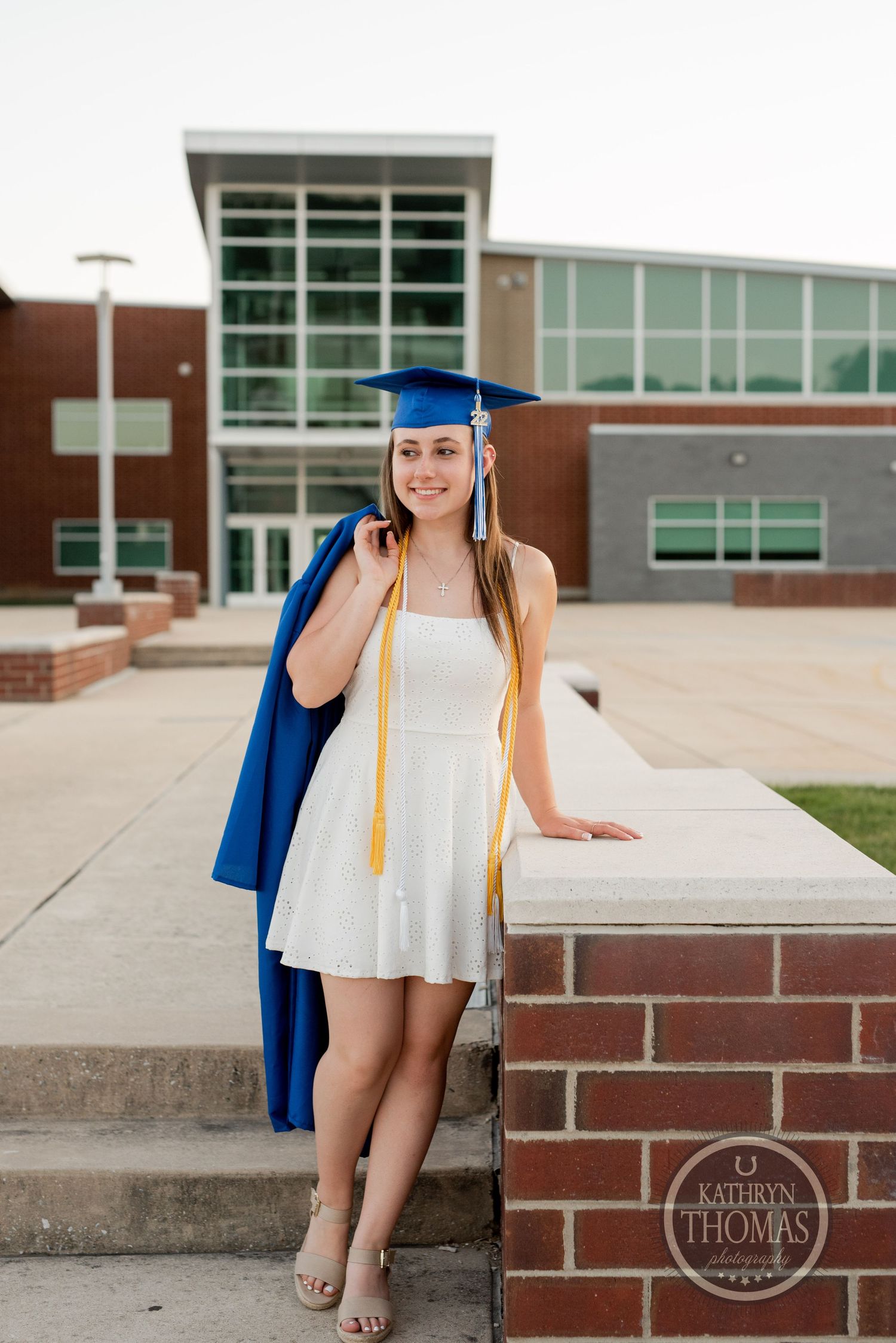 Graduate in a white dress and blue cap and gown poses in front of a modern brick school building.