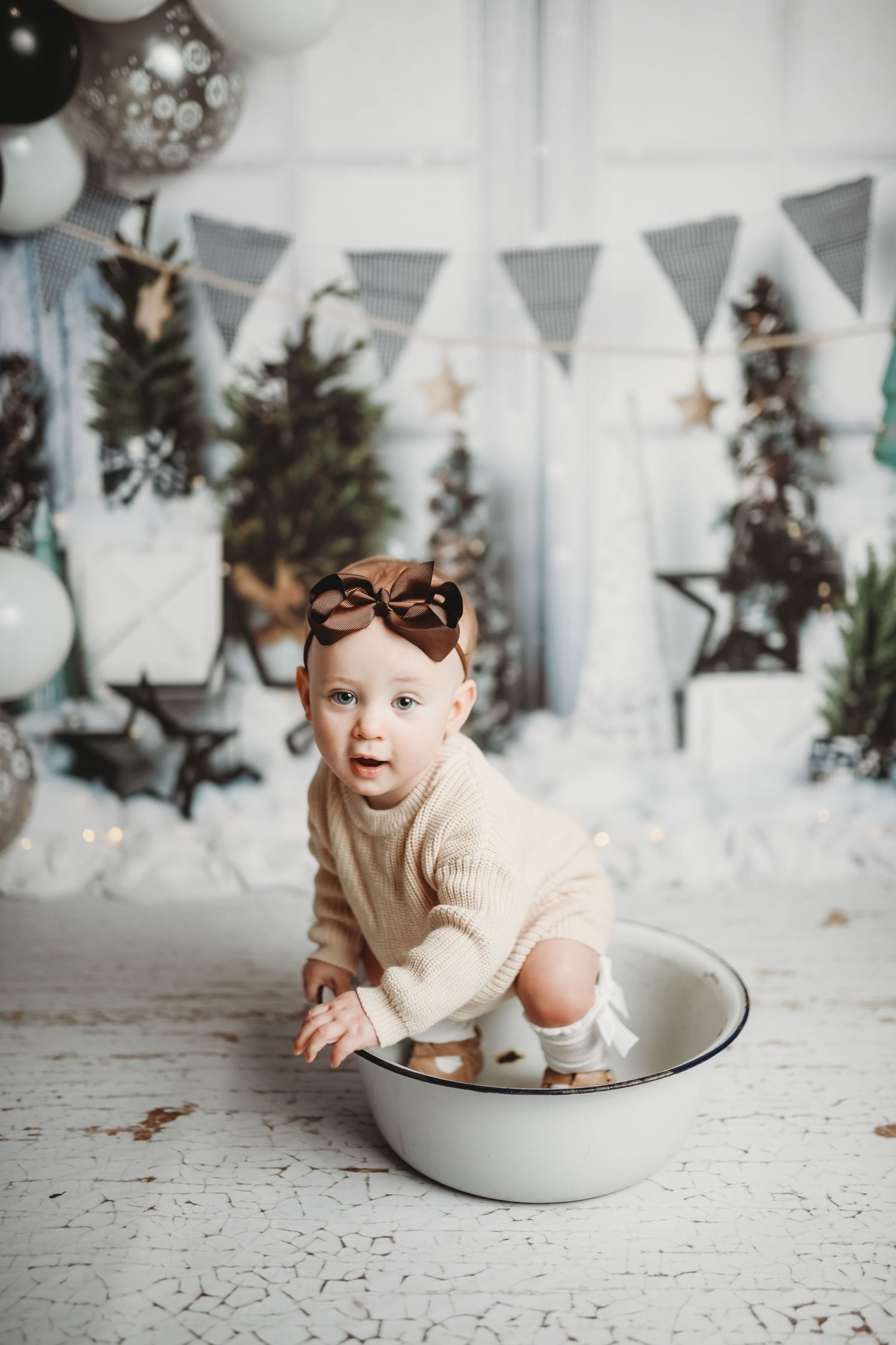 White winter themed setup with baby sitting in enamel bowl surrounded by holiday decorations.