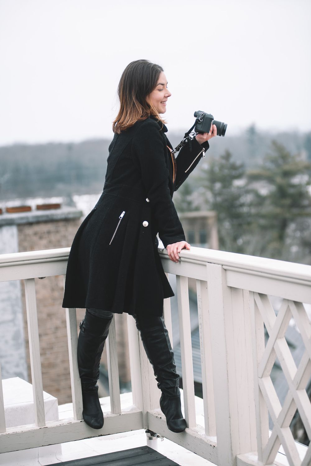 Laughing woman in black coat and boots holds camera on a white railed outdoor balcony.