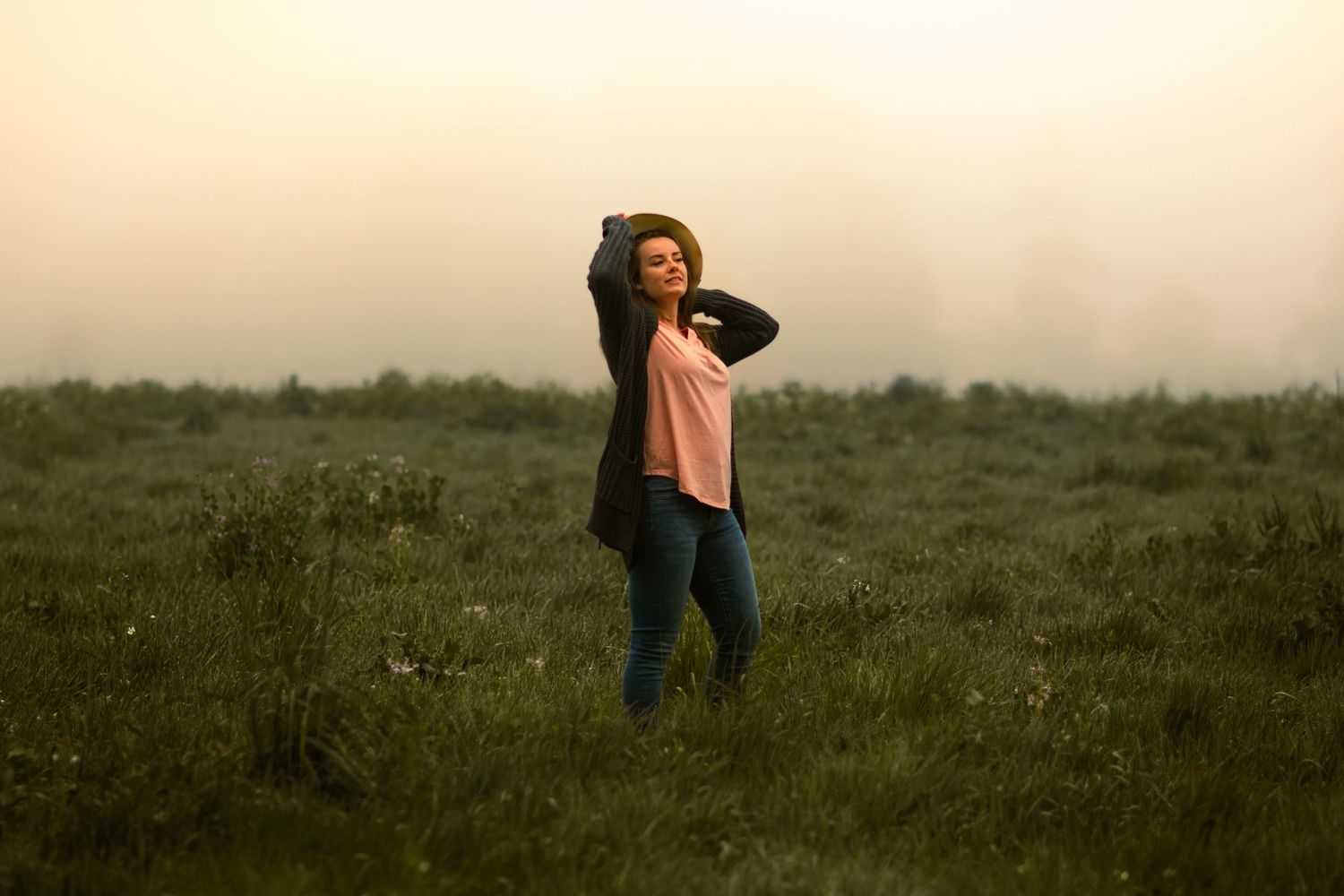 Ciara standing in field posing for picture. holding hat and looking off in the distance. Wearing pink shirt, skinny jean