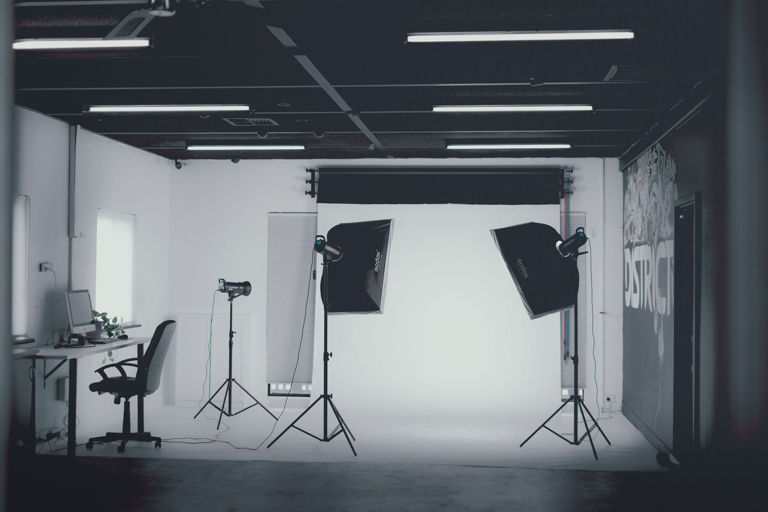 Professional photography studio with softbox lights on stands illuminating a white backdrop in a spacious industrial space.