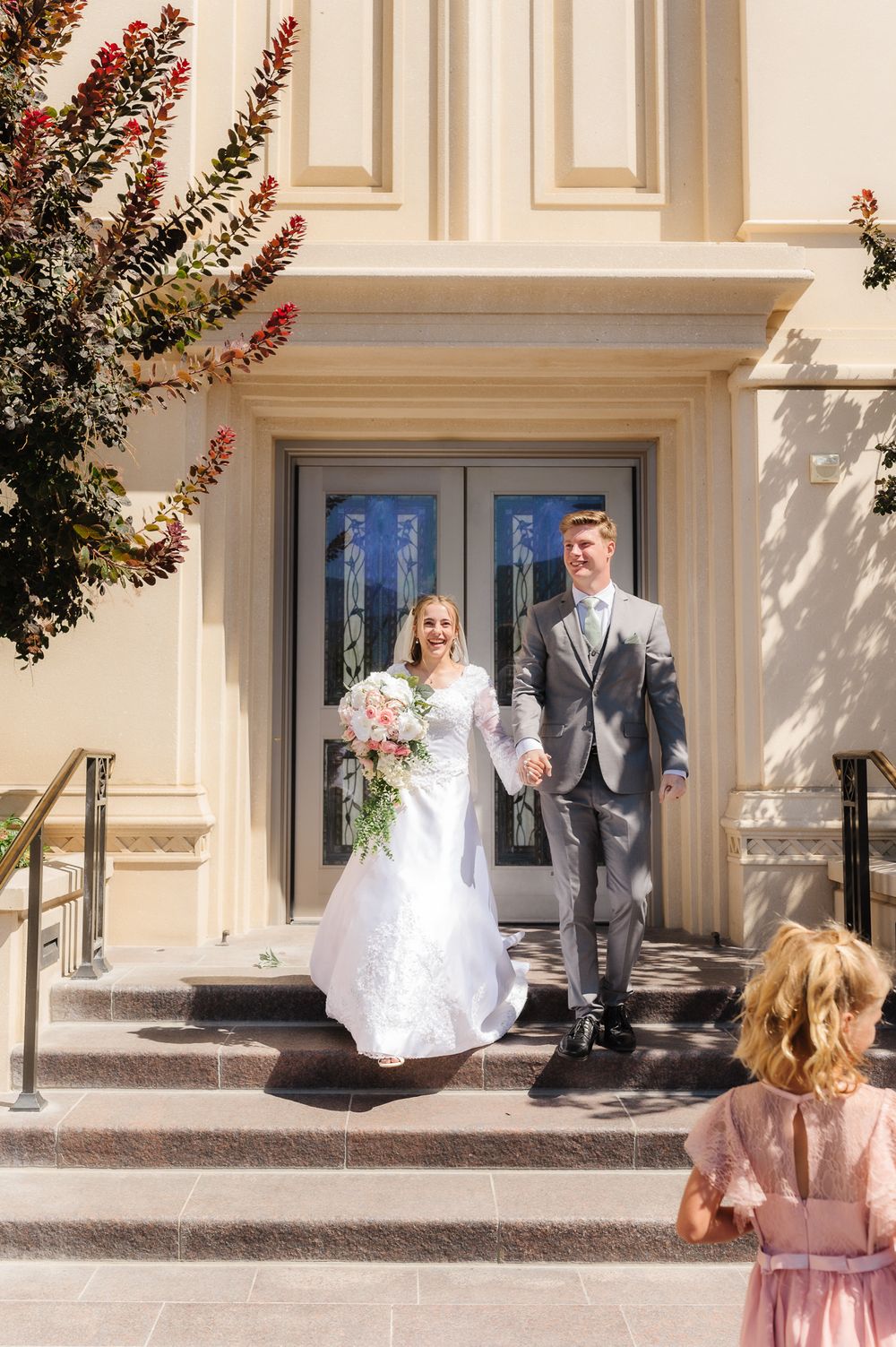 Wedding couple exits through ornate doors of a classic white building with decorative trim.
