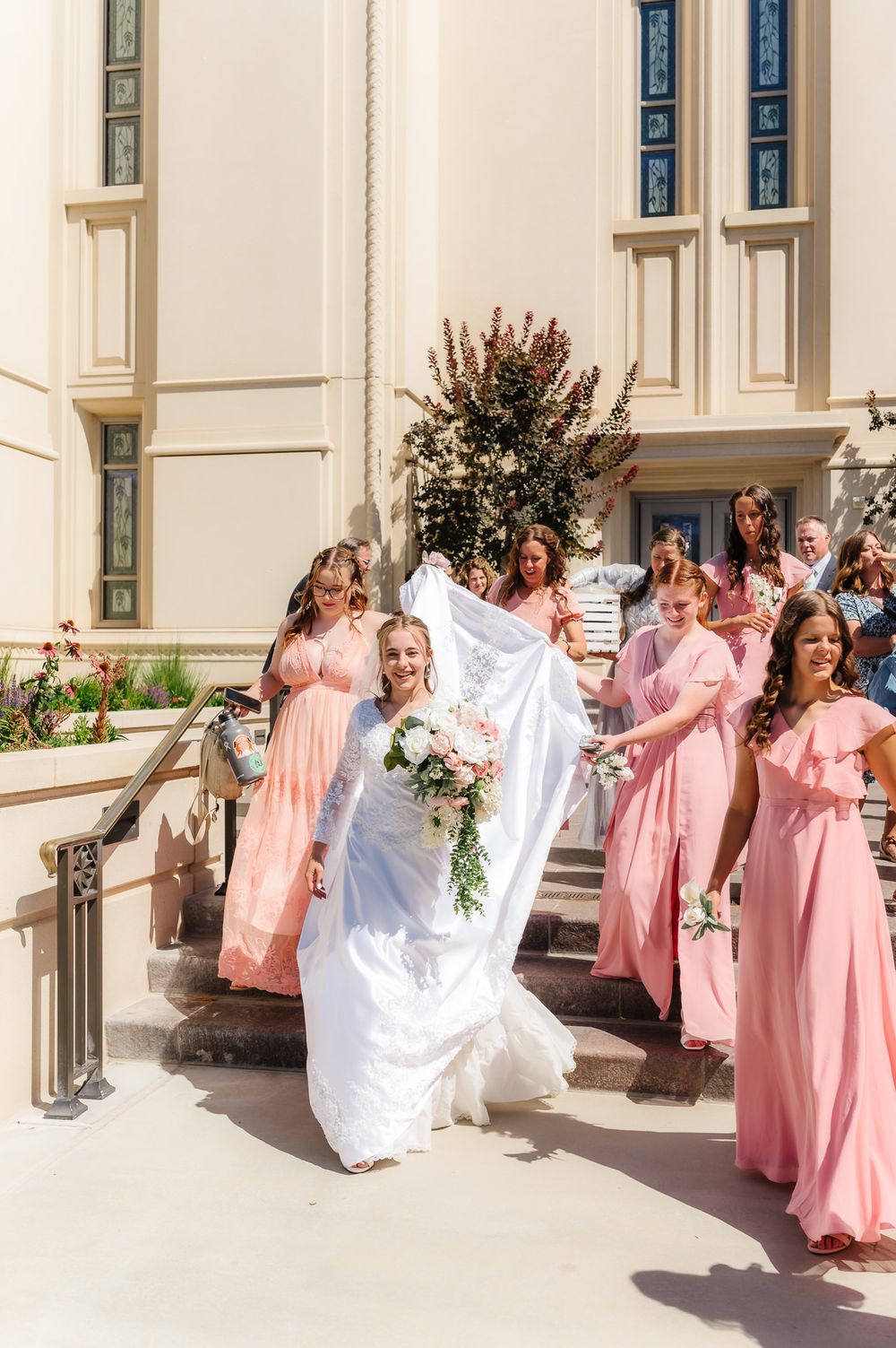 Wedding party members in pink dresses celebrate joyfully outside a grand white building.