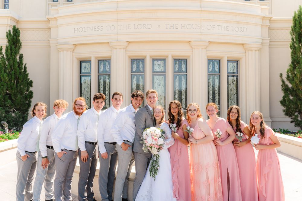 Wedding party in pink dresses and gray suits poses in front of white stone building.