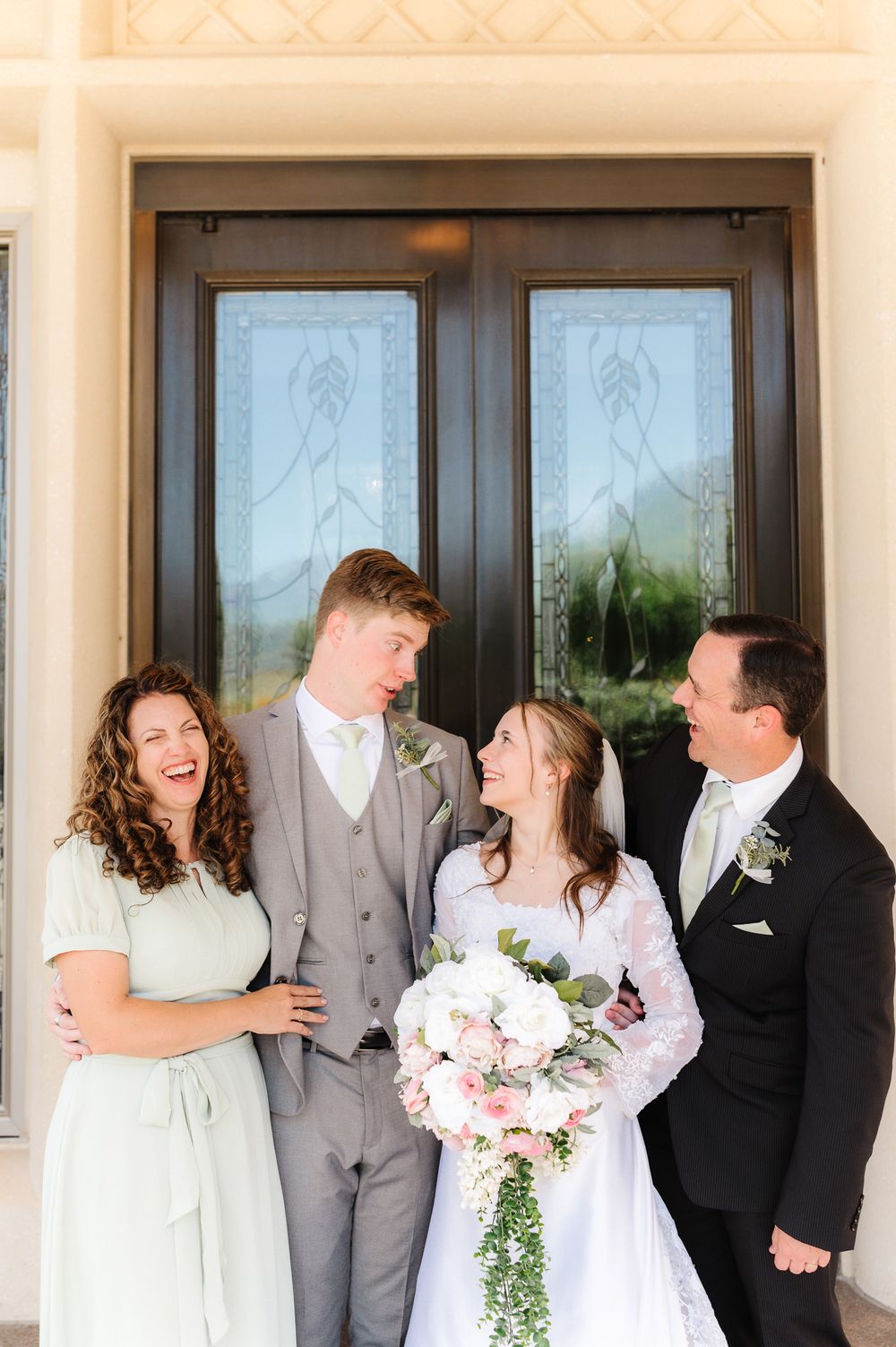 Wedding party poses casually in a line outside venue with bride in white and groomsmen in gray suits.