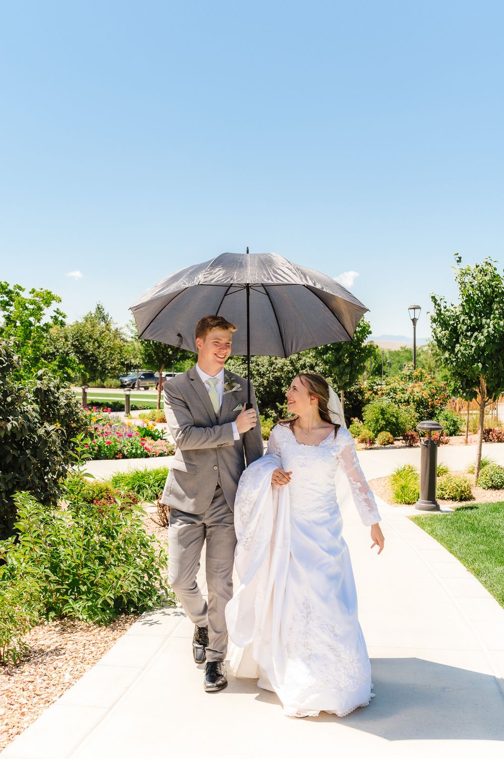 Wedding couple walking with a silver umbrella in a sunny garden setting.