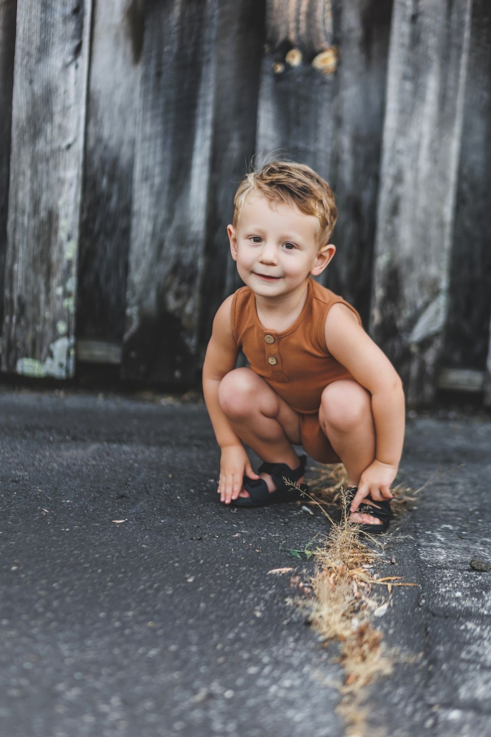 Candid portrait of young child crouching near weathered wood wall.
