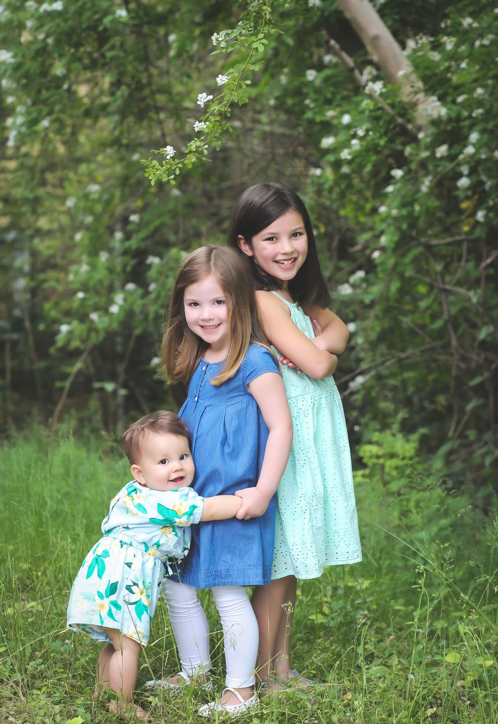Three siblings pose together in pretty dresses among blooming trees.