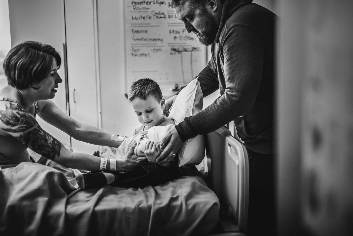 A young boy is handed his newborn brother to hold for the first time as his sits on a hospital bed between his folks.