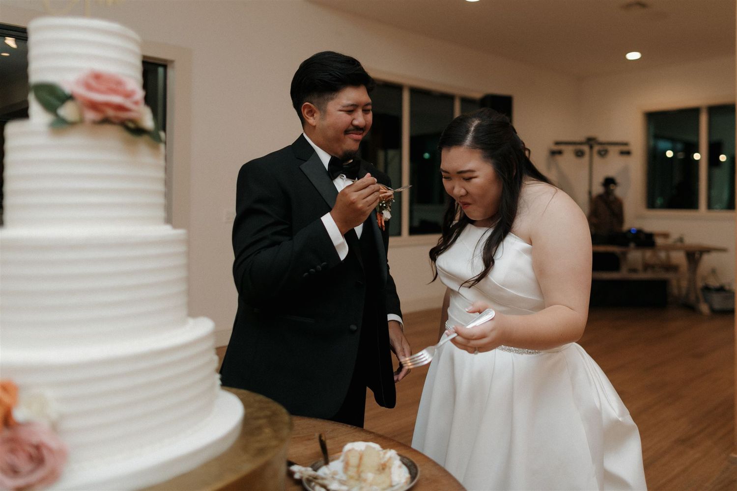 Couple sharing cake during wedding reception beside tiered white cake.