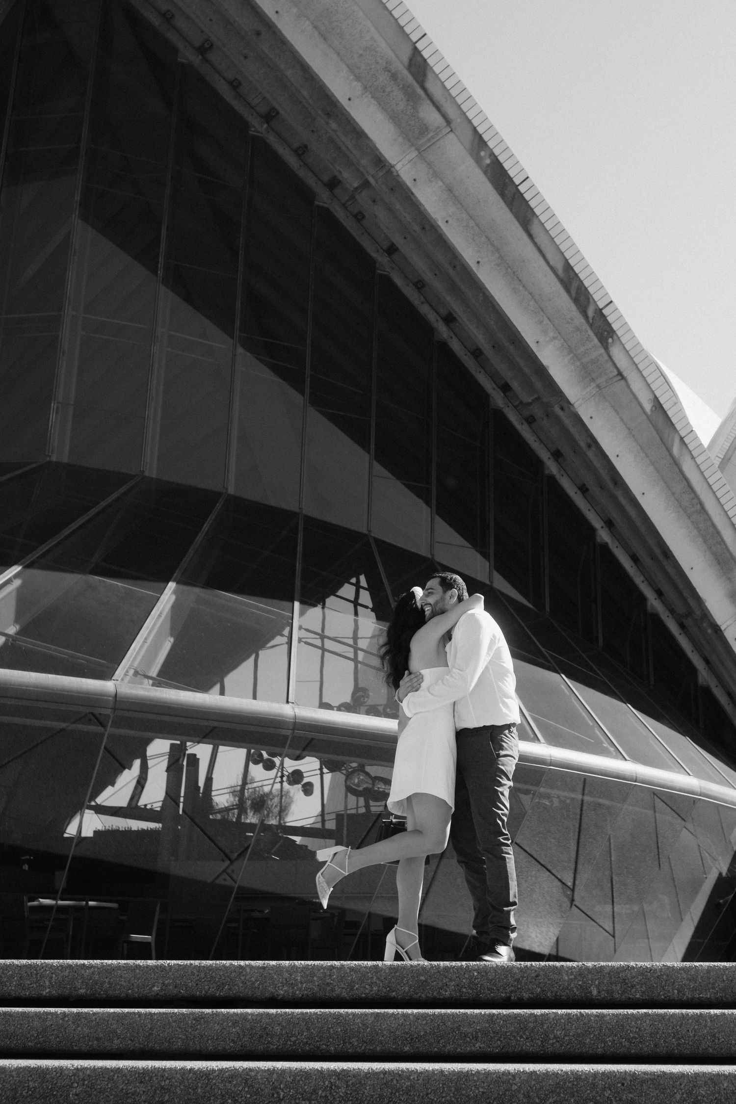 A romantic black and white moment captured under the dramatic architecture of the Sydney Opera House.