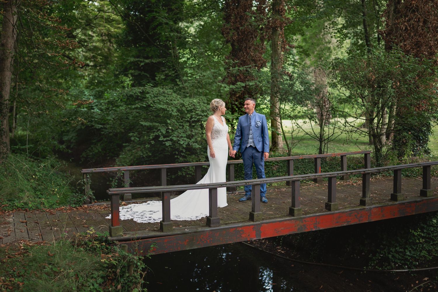 A couple stands on a wooden bridge surrounded by lush greenery for their wedding photos.