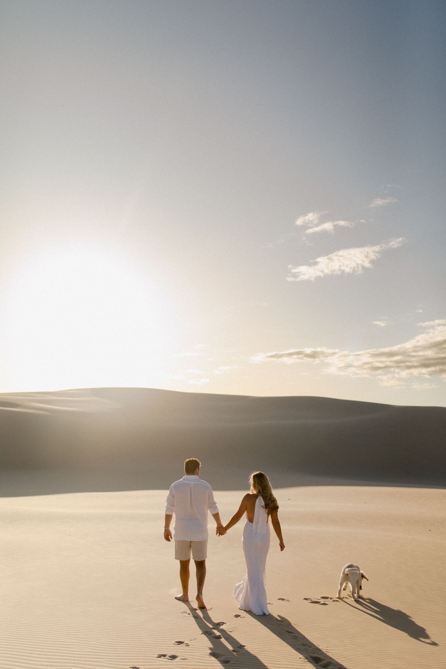 Couple walking hand in hand across sand dunes with dog at sunset.