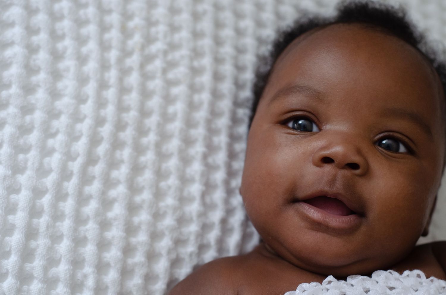Adorable smiling Black baby lying on a white textured blanket looking up at the camera.