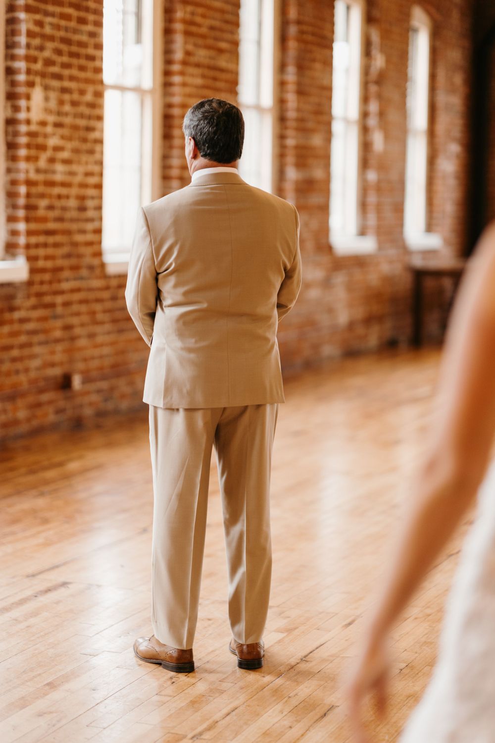 Father of the bride in tan suit standing with back turned awaiting first look on wood floor.