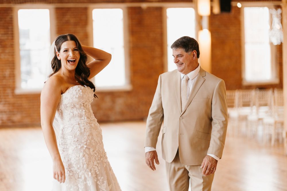 Bride laughs joyfully with groom in tan suit during first look at rustic brick wedding venue.
