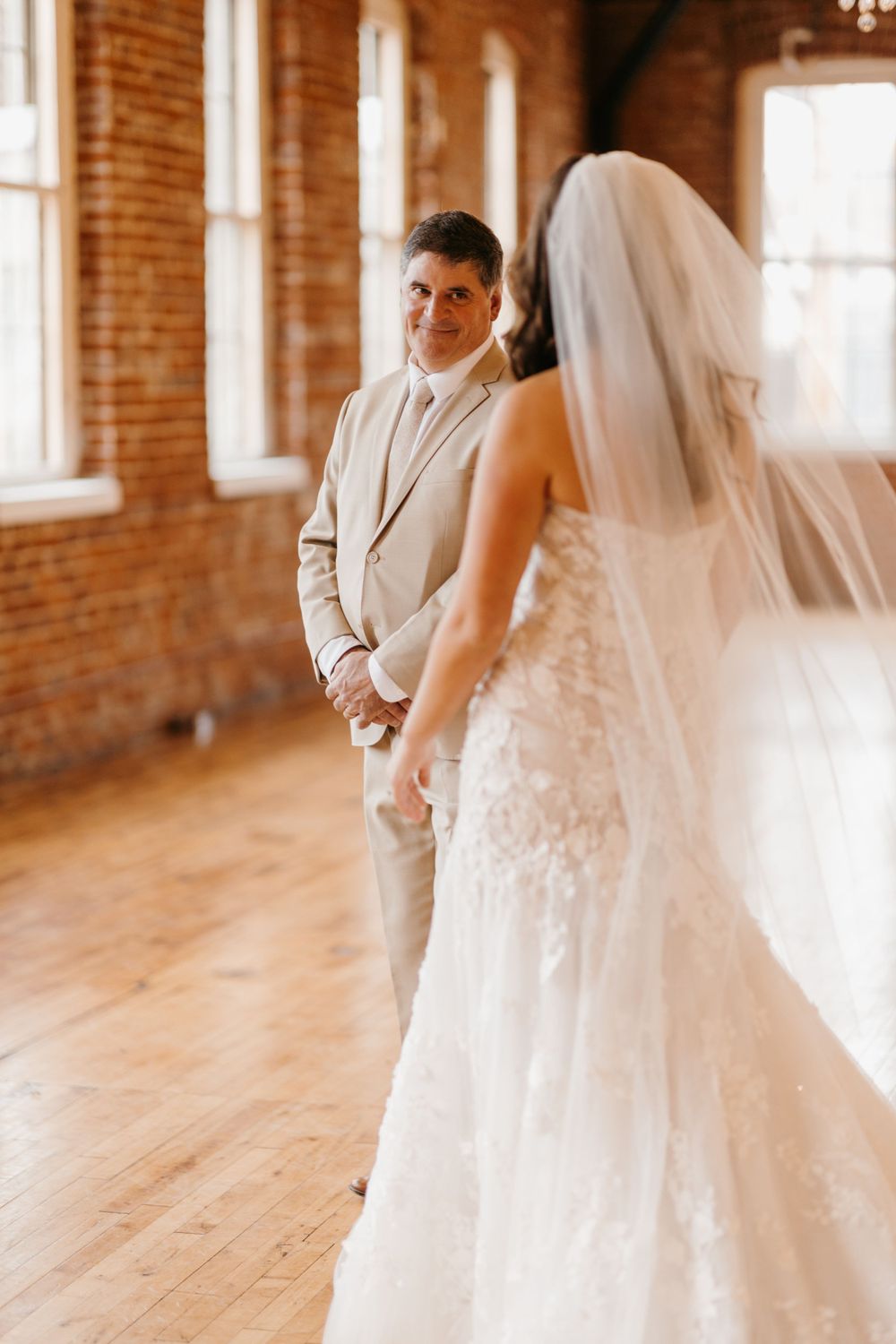 Groom in tan suit smiles at bride in lace wedding dress during first look in brick venue.