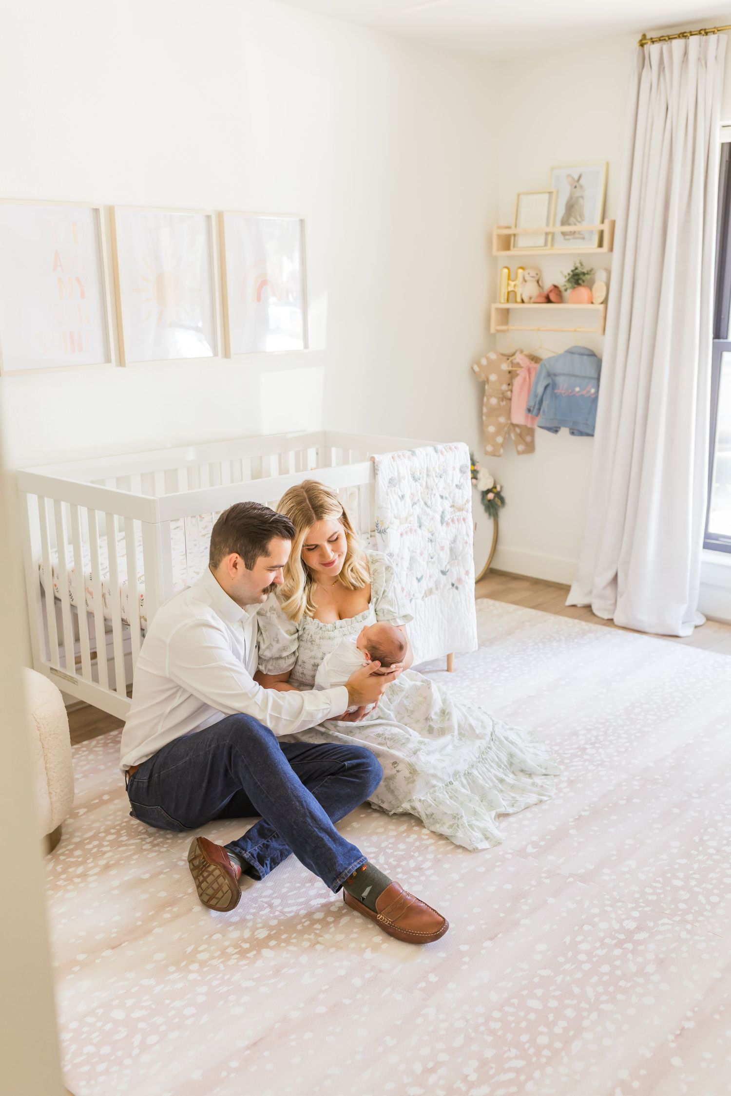 A couple sits on the nursery floor with their newborn baby near a white crib.