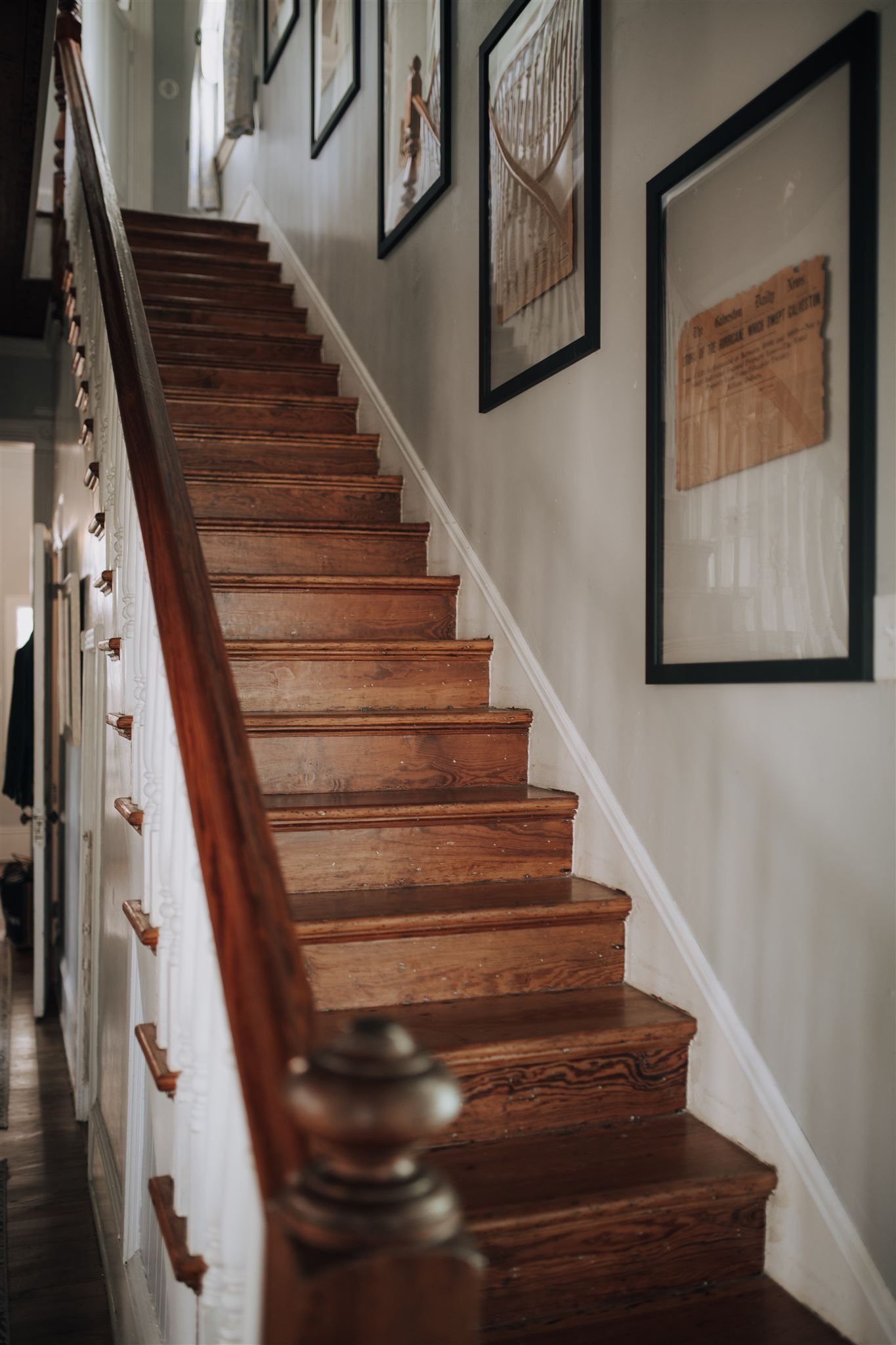Wooden staircase with white walls features framed artwork and ornate banister details.