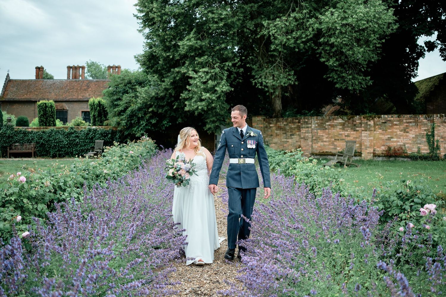 Couple strolls through blooming lavender garden with stone wall and buildings in background.