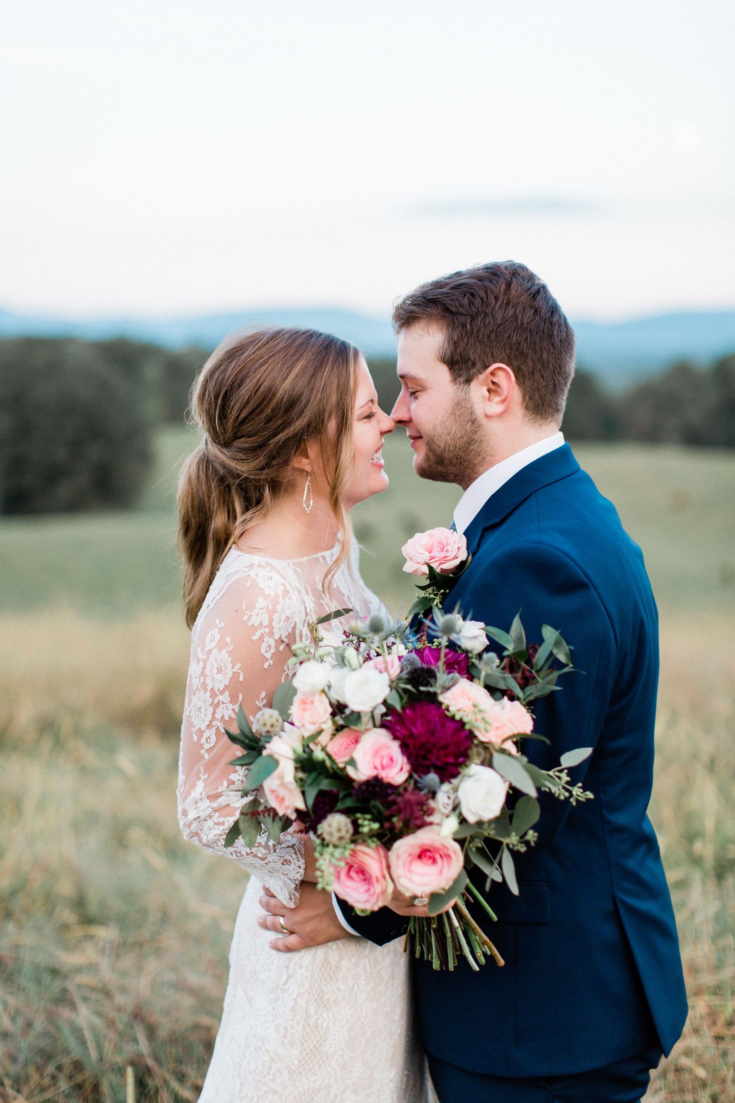 Bride and groom share romantic nose-to-nose moment in field, bride holding colorful bouquet.