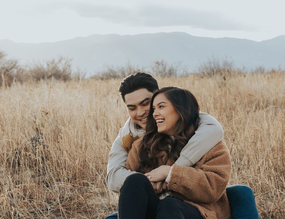 Couple laughing together while sitting in tall golden grass with mountains in background.