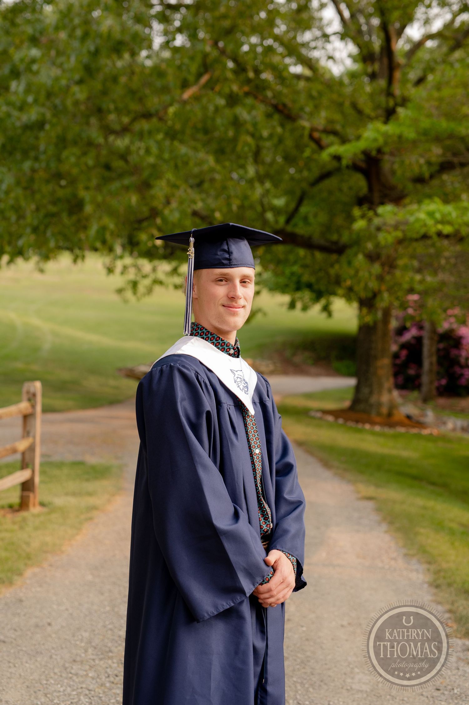 Graduate in navy blue cap and gown poses on a scenic path with trees in the background.