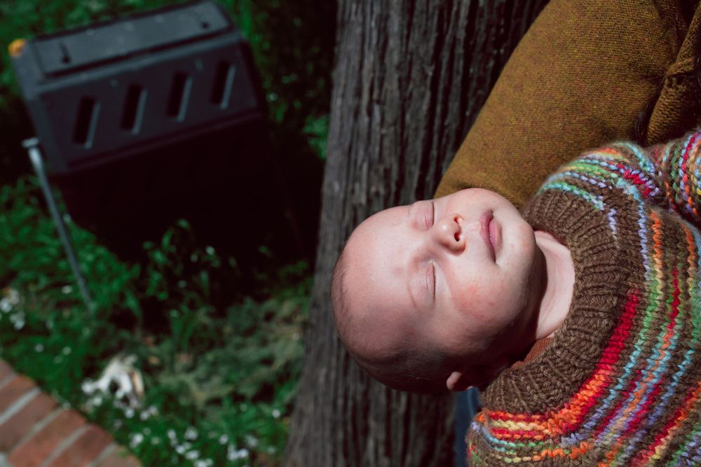 Child in colorful striped sweater resting against a tree trunk in a garden setting.