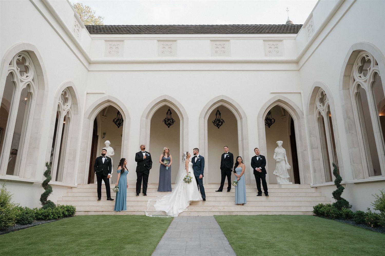A wedding party stands in an elegant colonnade courtyard with Gothic arches and landscaped gardens.