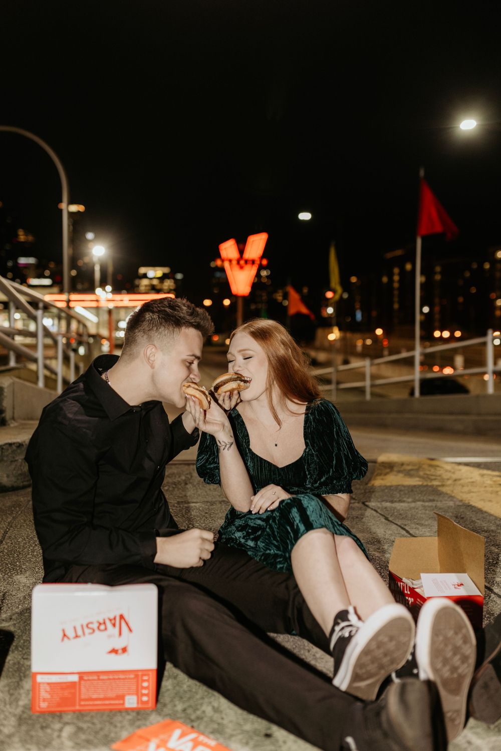 A couple sharing food at night with a bright neon sign glowing in the background.