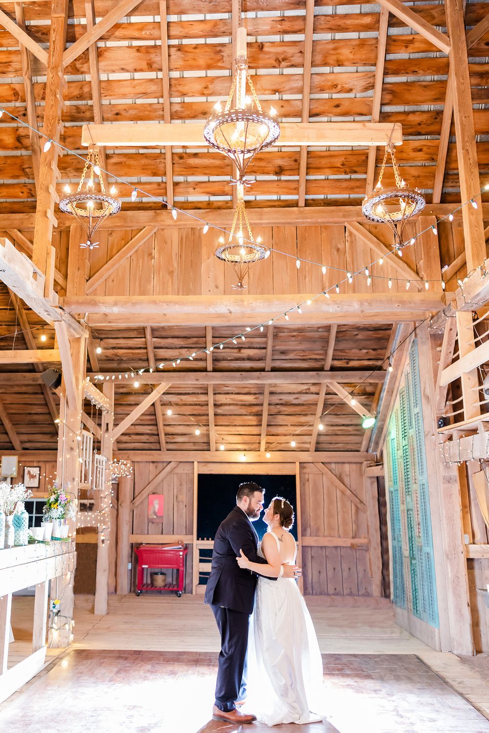 Newlyweds embrace under crystal chandeliers in a rustic wooden barn venue with twinkling string lights.