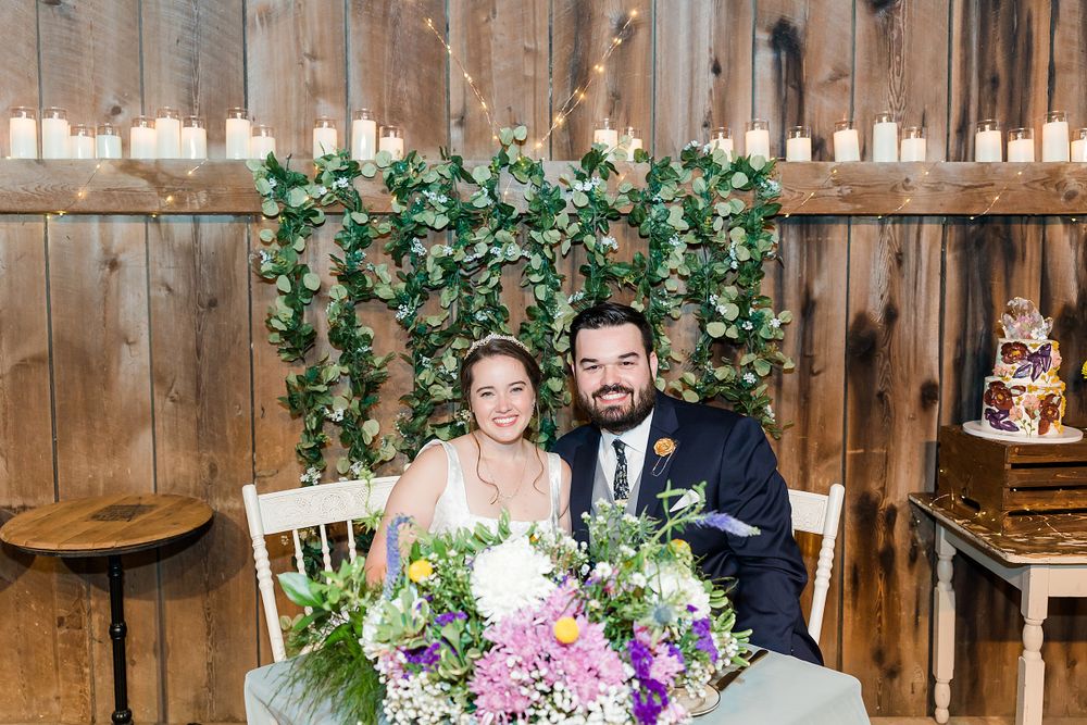 Couple seated at rustic barn wedding reception with floral decorations and wooden backdrop.