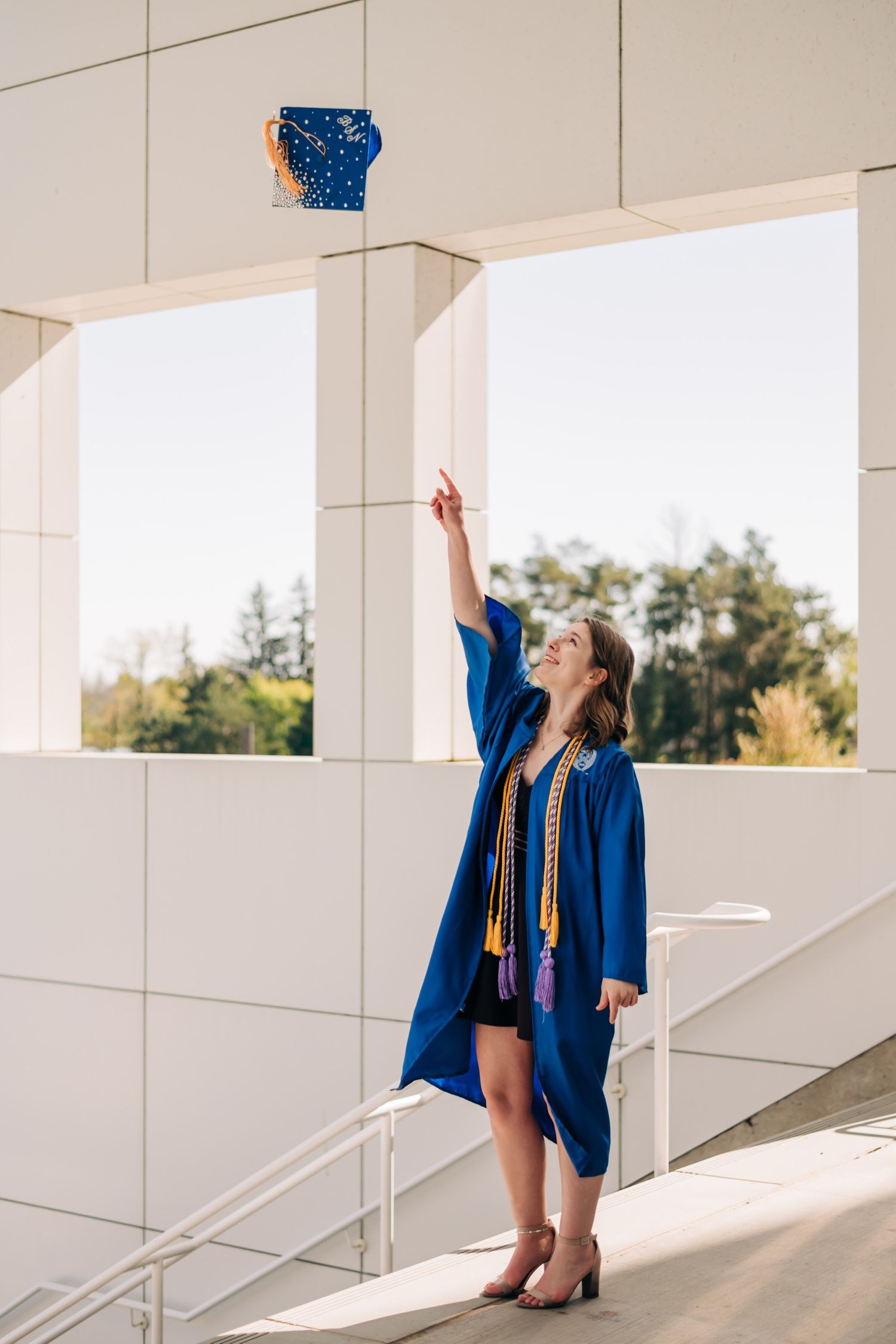 female college graduate wearing blue graduation gown with yellow cords, ceremoniously throwing blue cap into the air