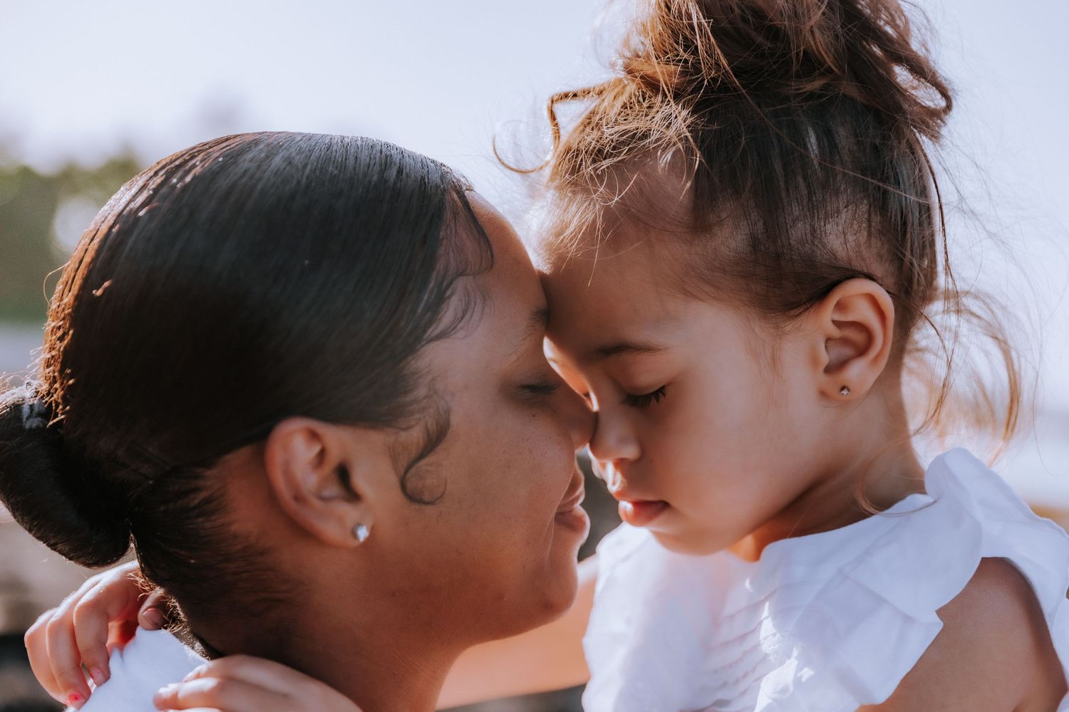 Tender moment between parent and child with faces close together against bright background.