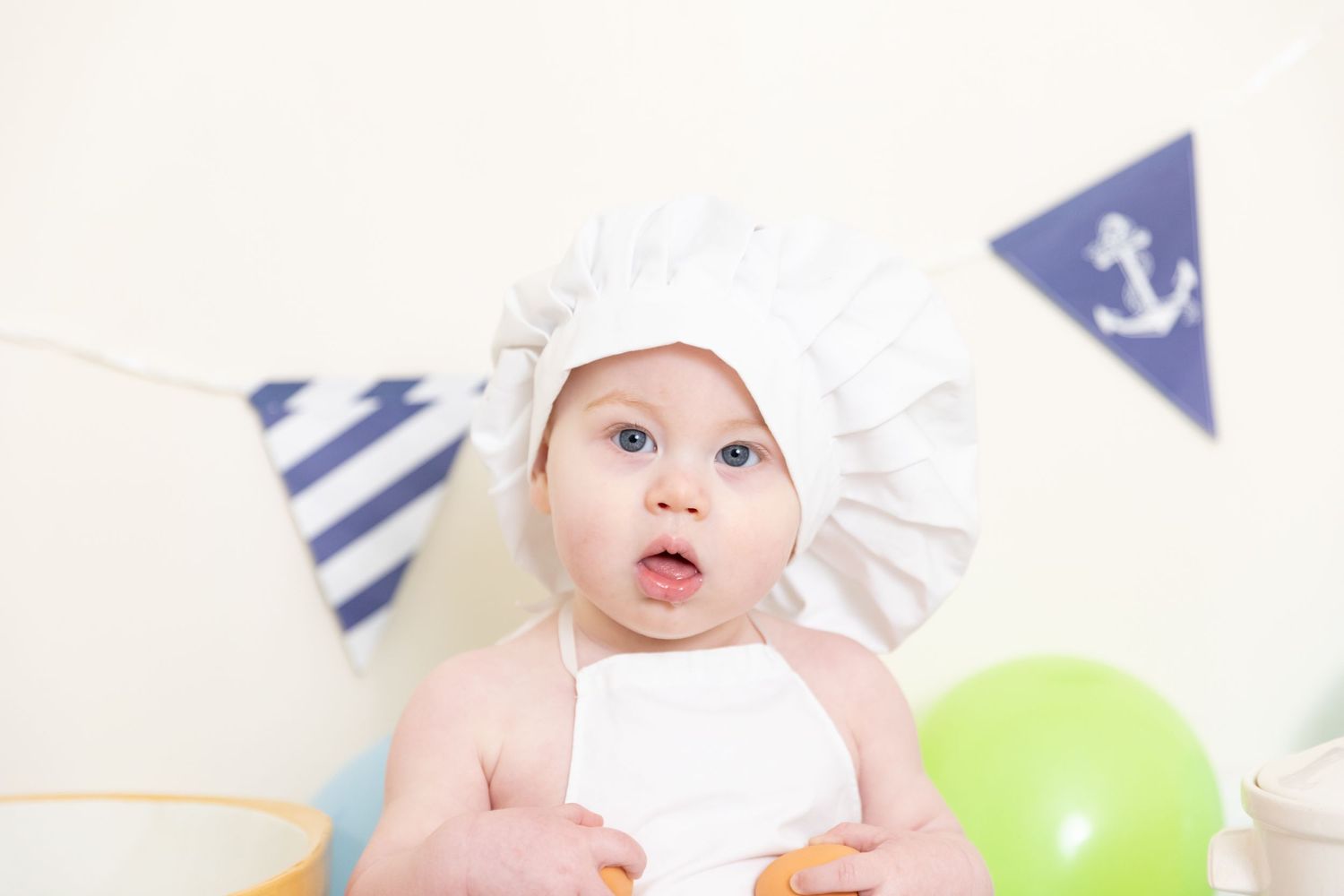 A baby in a white bonnet with nautical-themed party decorations and balloons.