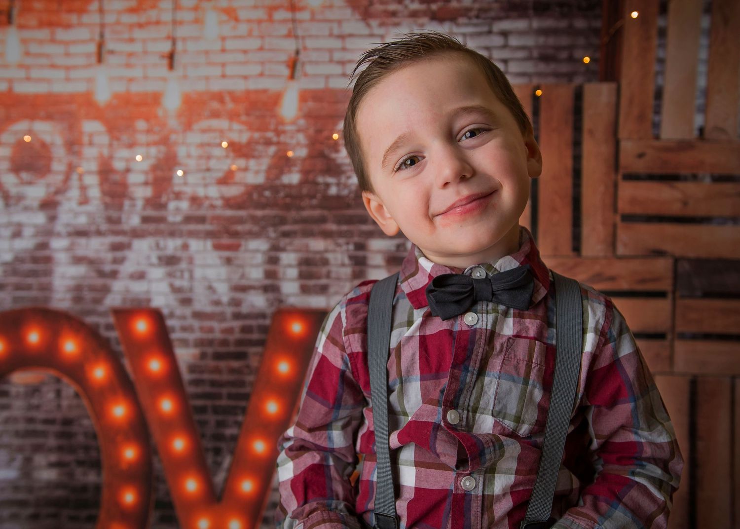 A child in plaid and suspenders poses in front of a rustic brick wall with illuminated letters.