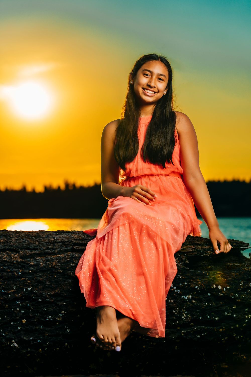 Person sits on rock at sunset wearing coral dress against vibrant sky and water.