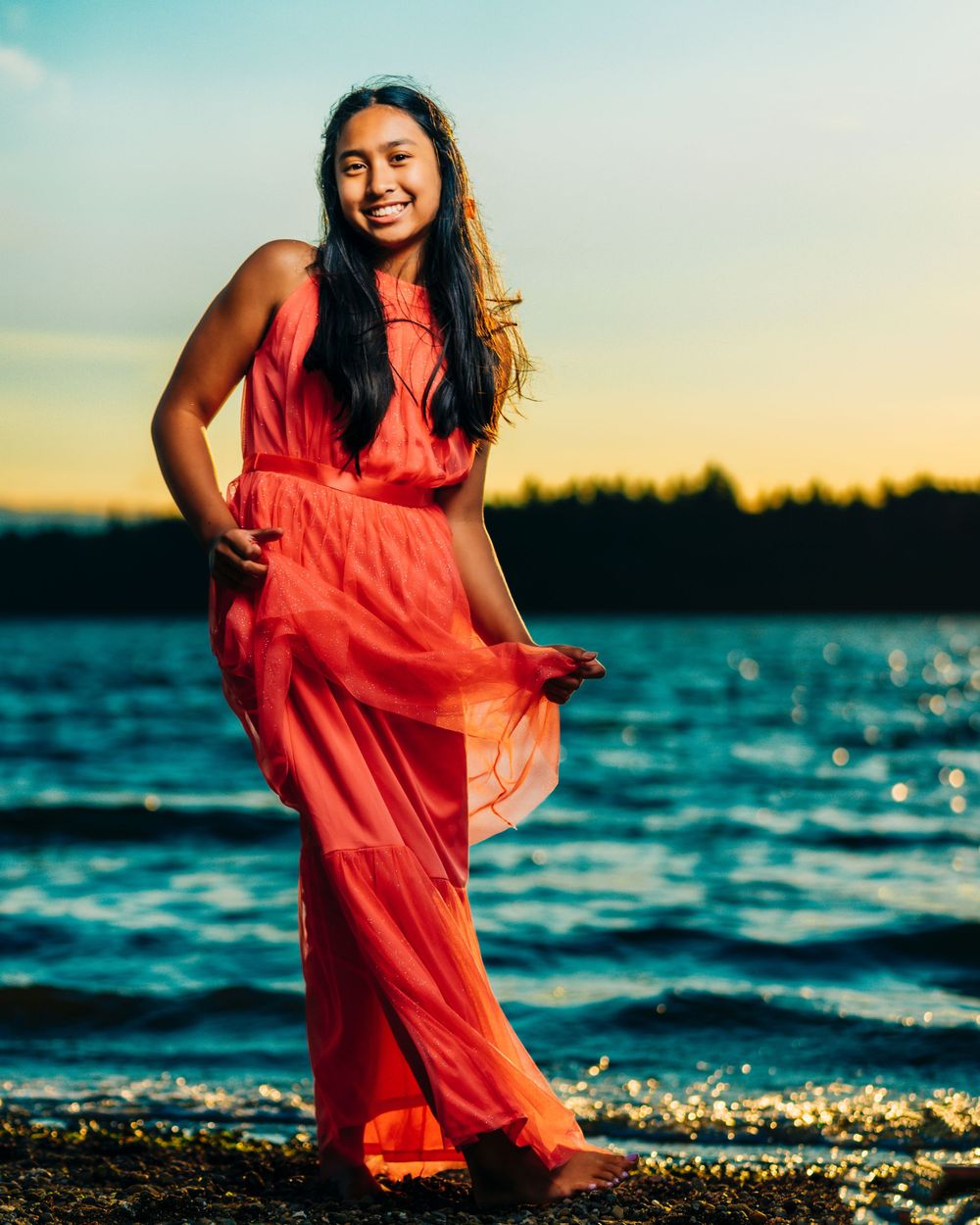 Person in flowing coral dress poses by water during golden hour with sparkling waves.
