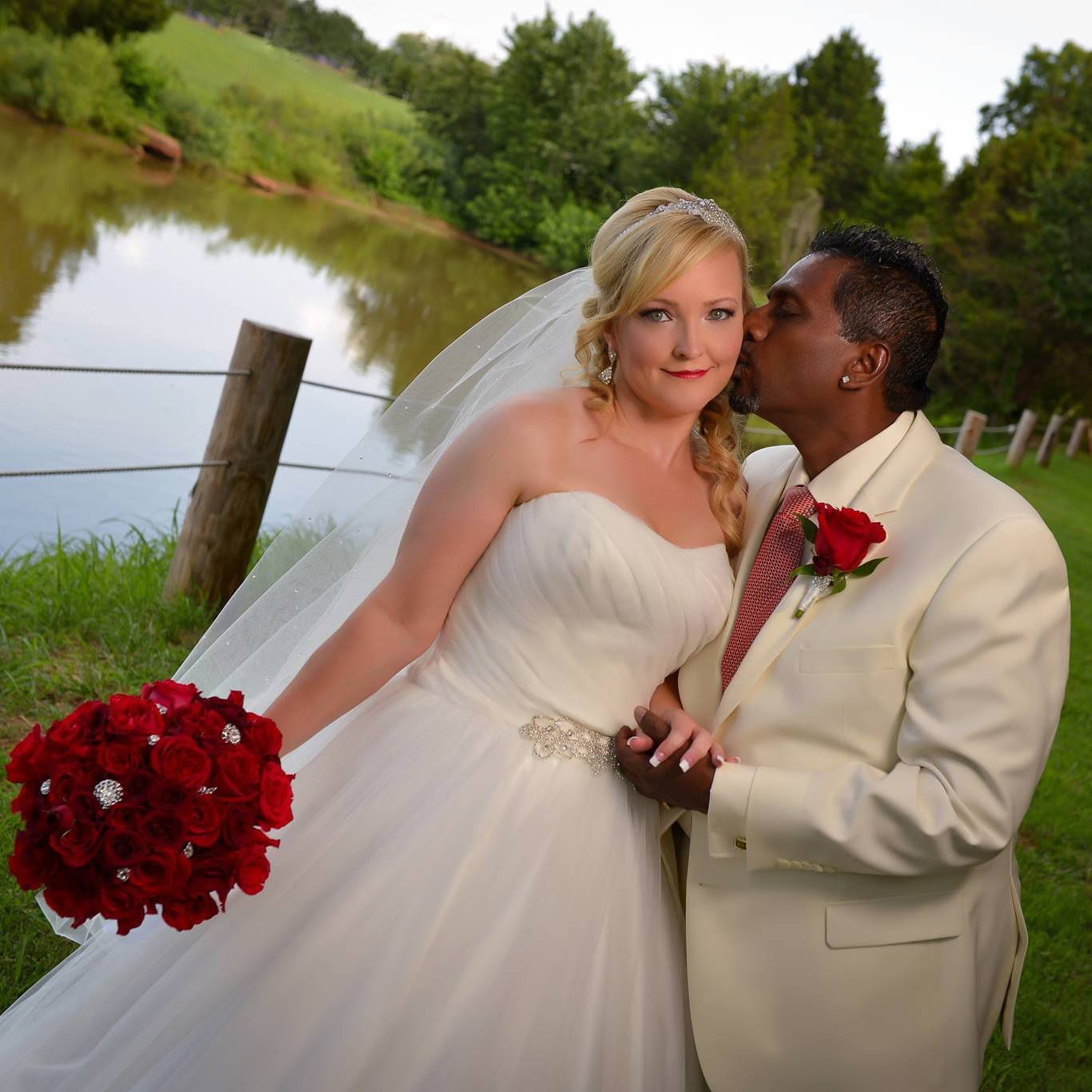 A couple shares romantic moments by a lake with red roses and formal attire.