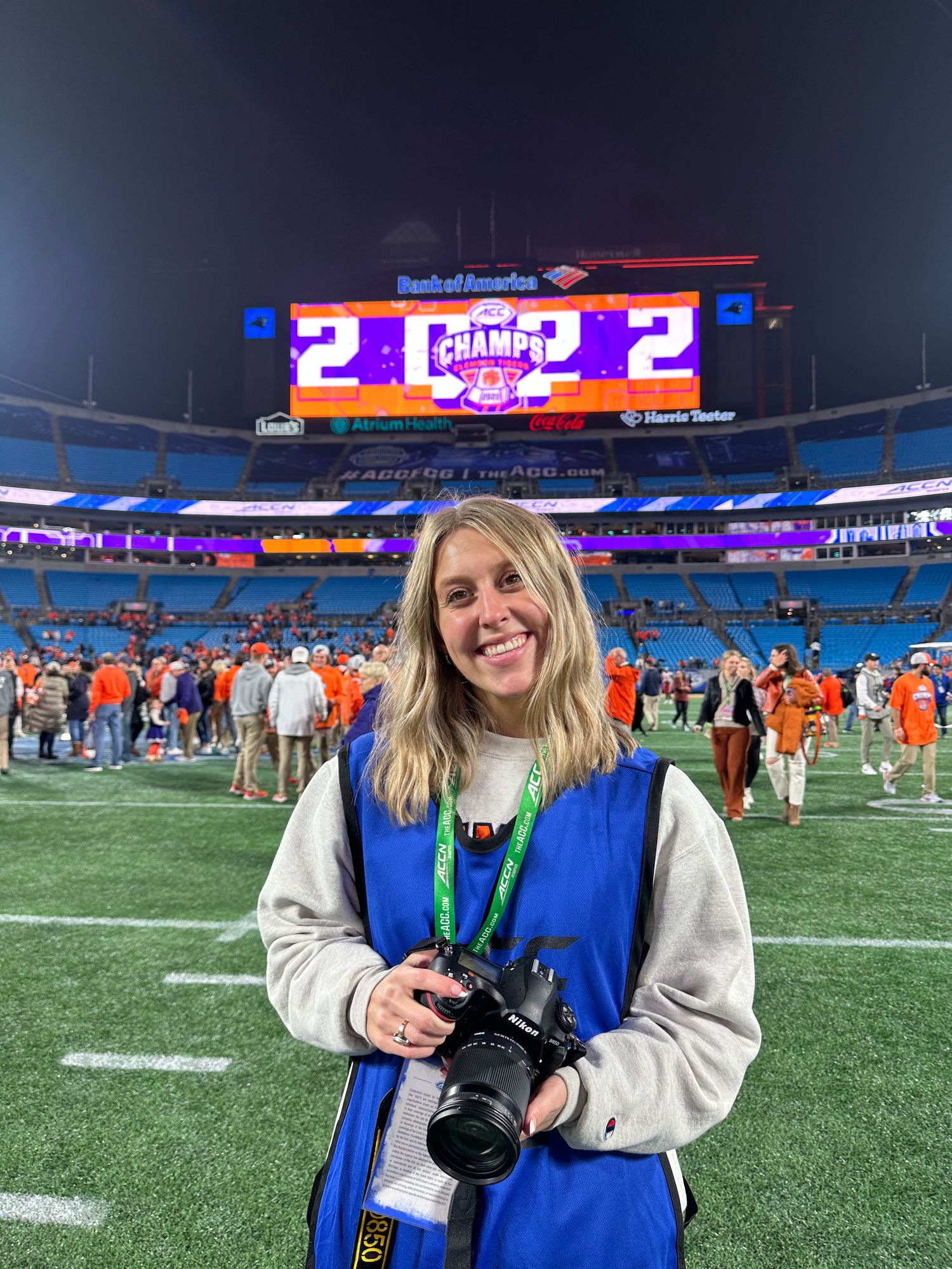 Photographer with camera standing on football field at night with illuminated scoreboard in background.