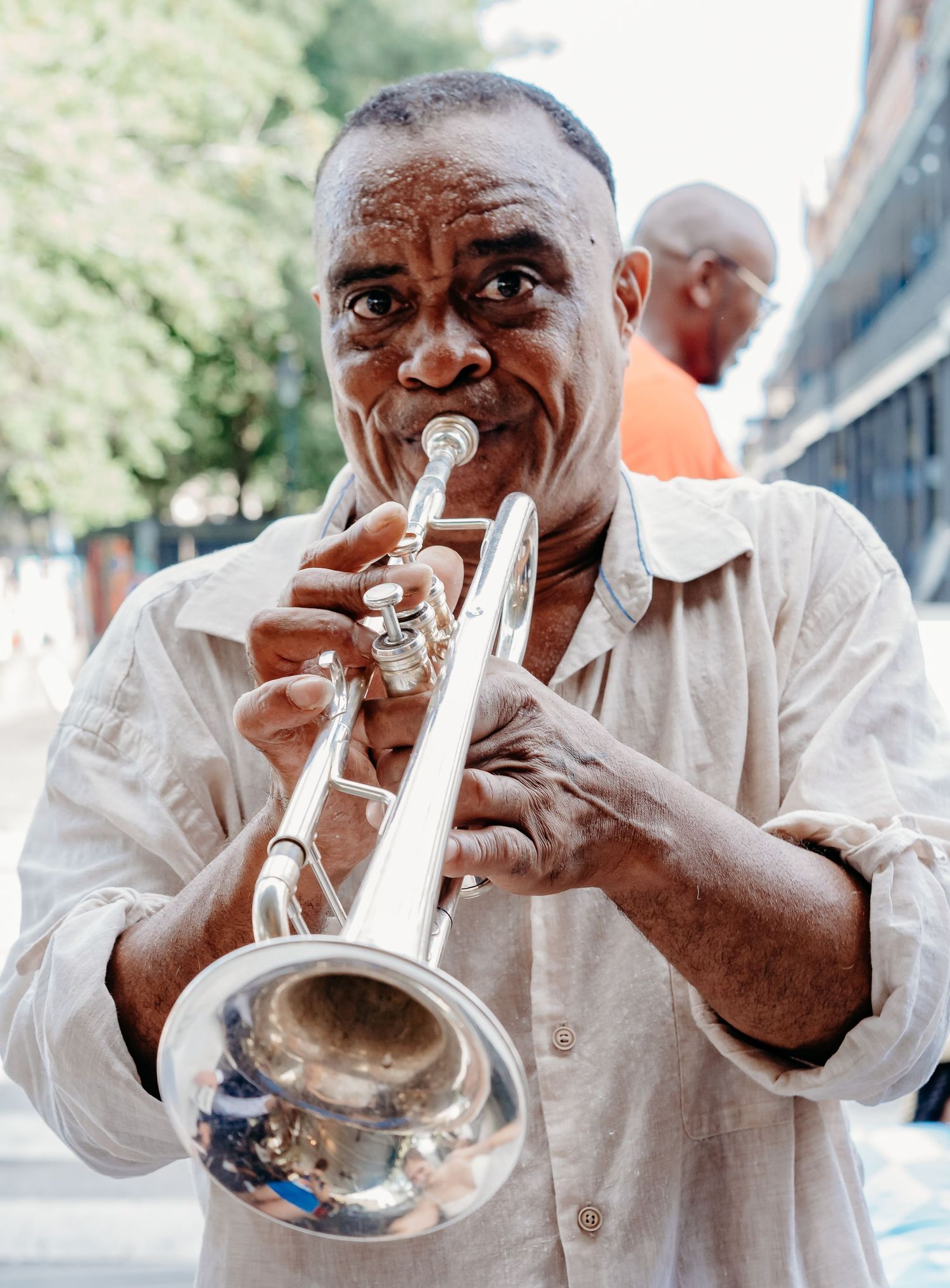 A musician playing a silver trumpet outdoors, wearing a light colored shirt.