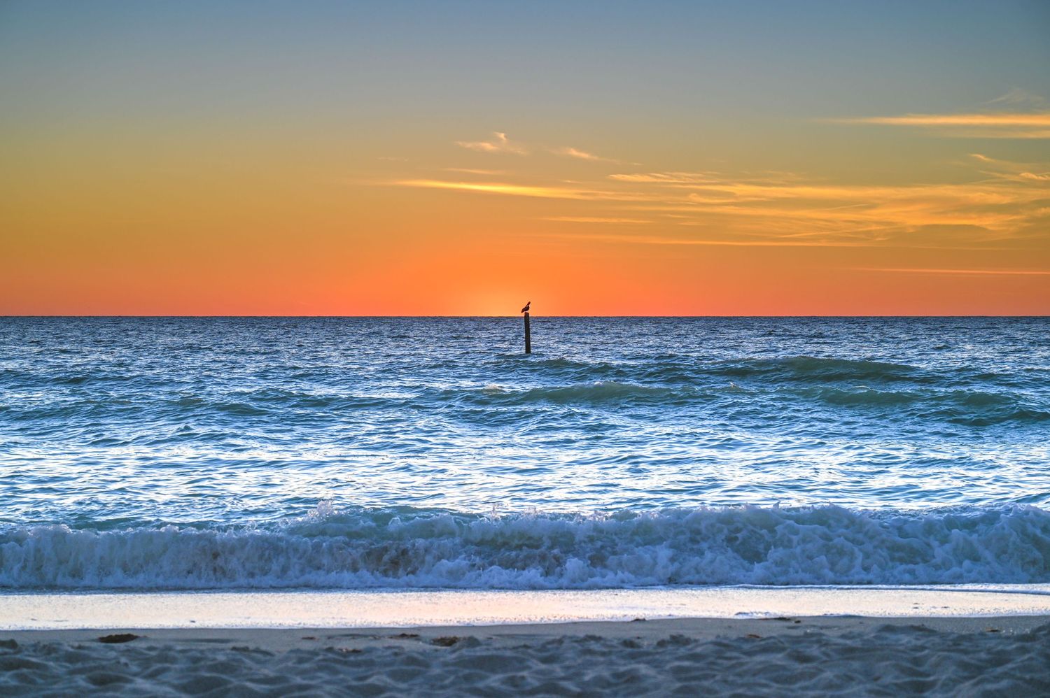 Orange and purple sunset over ocean waves with a distant lighthouse.