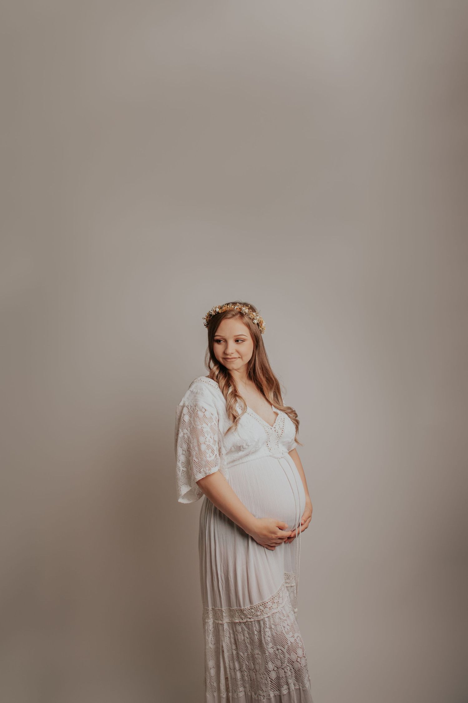 Maternity portrait of woman in white lace dress against neutral backdrop.