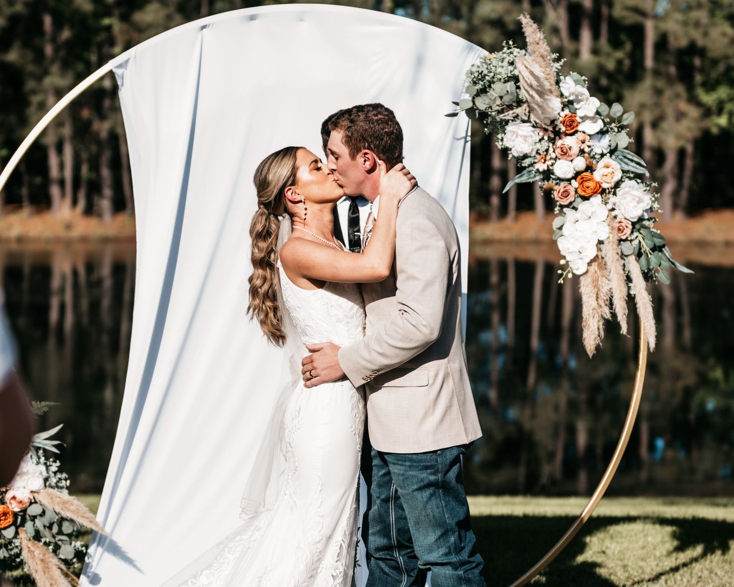 A romantic wedding moment captured under a circular arch with floral decorations beside a lake.
