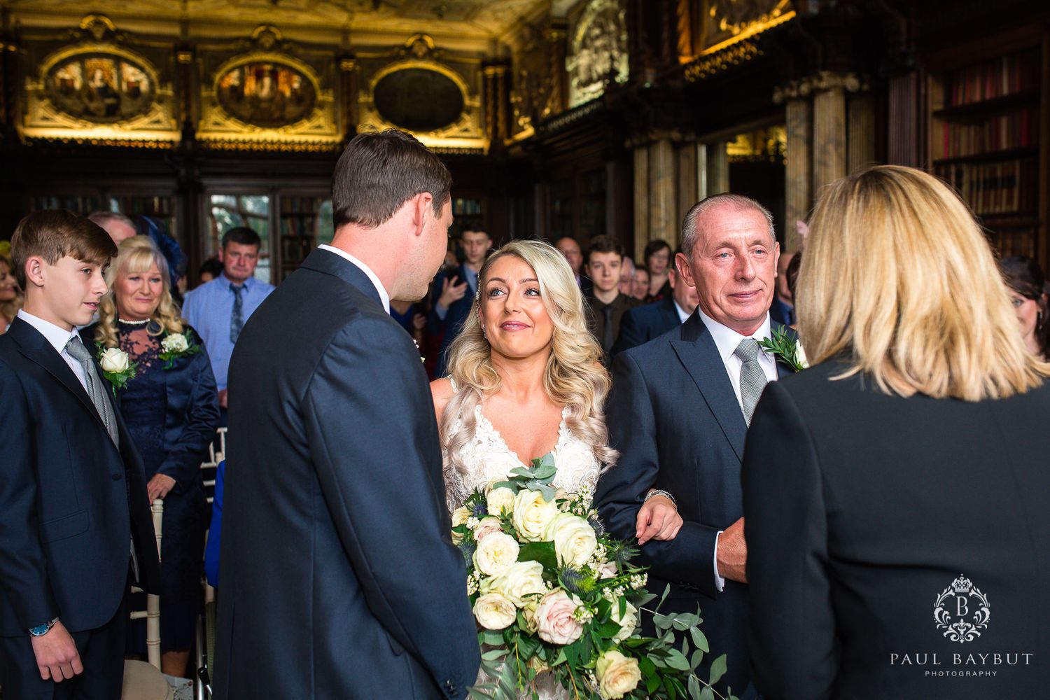 Wedding ceremony guests gather in an ornate historic venue with golden ceiling details.
