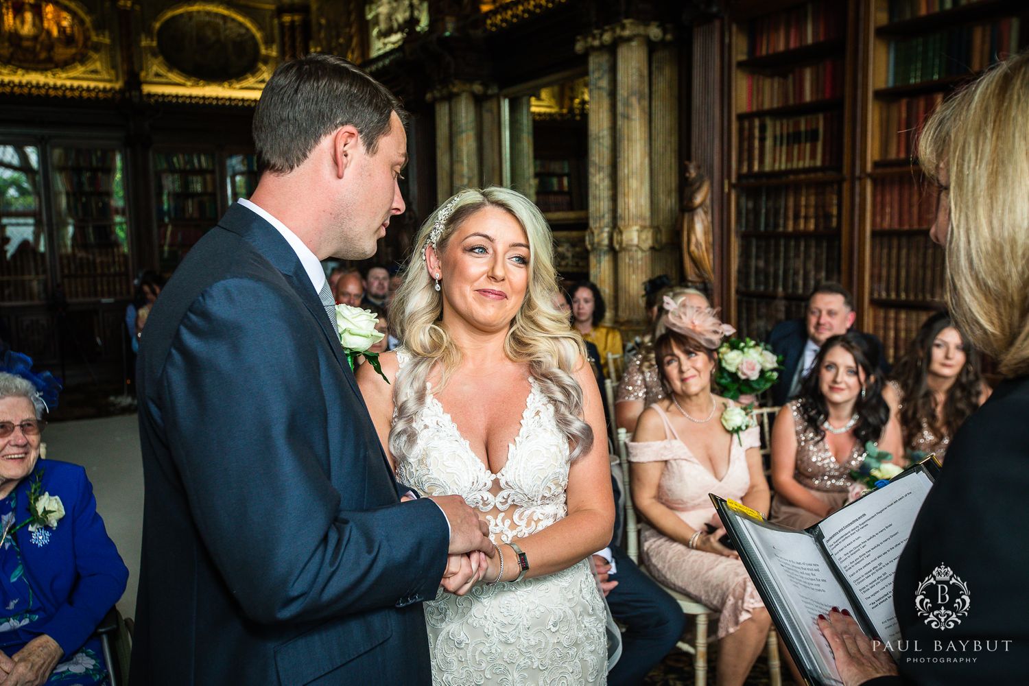A couple exchanges vows during an elegant wedding ceremony in an ornate library with guests watching.