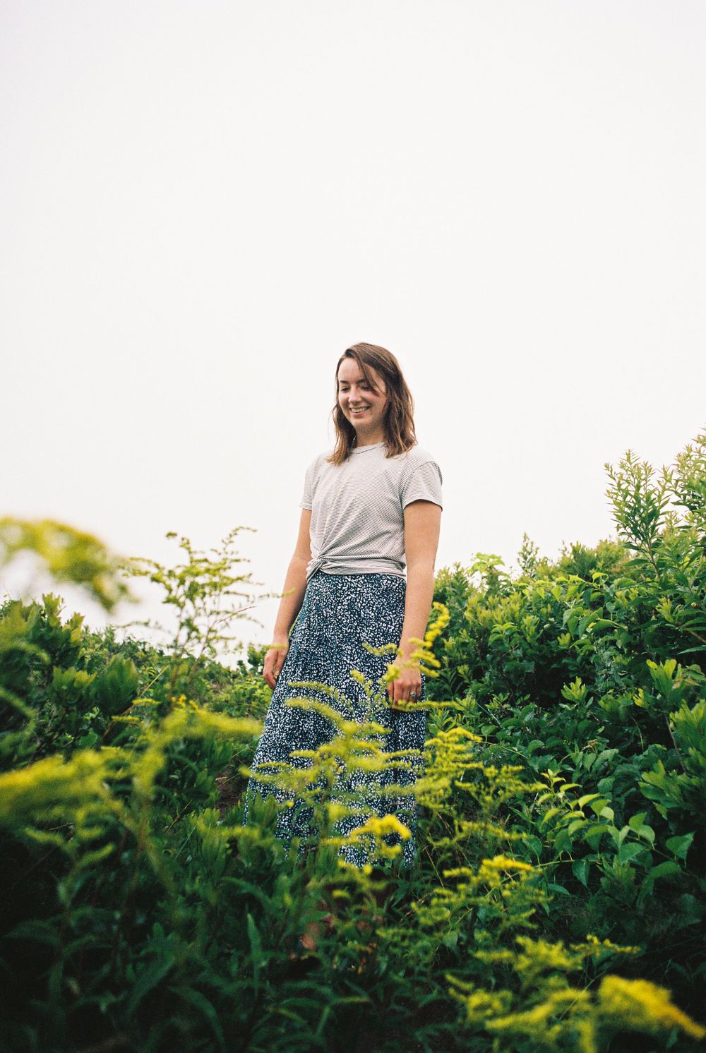 A person in a t-shirt and patterned skirt stands among lush summer foliage and yellow flowers.